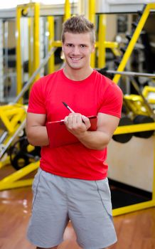 Young male personal trainer writing on clipboard, with training equipment behind him in gym