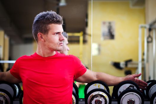 Handsome young man in gym sitting on dumbbells rack, looking to a side