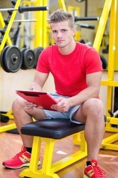 Young male personal trainer holding clipboard, sitting on training equipment in gym