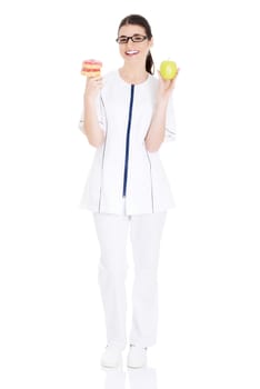 Young female doctor, nurse holding an apple and doughtnuts. Isolated on white.