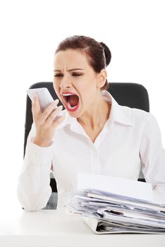 Beautiful business woman sitting, screaming to a phone and has a stack of papers on desk. Isolated on white.