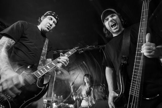 Photo of a band playing on a stage. Male guitarist, male bassist and female drummer. Shot with strobes and slow shutter speed to create lighting atmosphere and blur effects. Slight motion blur on performers.