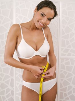 Photo of a slim young woman in her underwear measuring her waist with a tape measure.