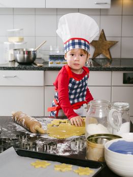 Photo of an adorable boy in a chef hat and apron making snowflake cookies in the kitchen.