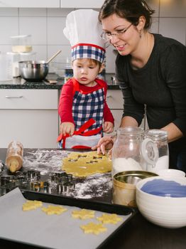 Photo of an adorable boy in a chef hat and apron and his mother making cookies in the kitchen.