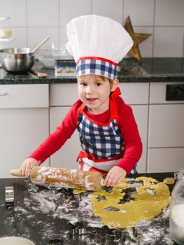 Photo of an adorable boy in a chef hat and apron using a rolling pin and making cookies in the kitchen.