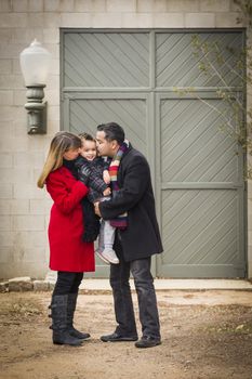 Young Mixed Race Couple in Winter Clothing Hugging and Kissing Son in Front of Rustic Building Together.