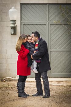 Young Mixed Race Couple in Winter Clothing Hugging and Kissing Son in Front of Rustic Building Together.