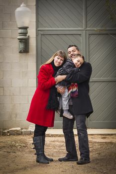 Young Mixed Race Couple in Winter Clothing Hugging and Kissing Son in Front of Rustic Building Together.