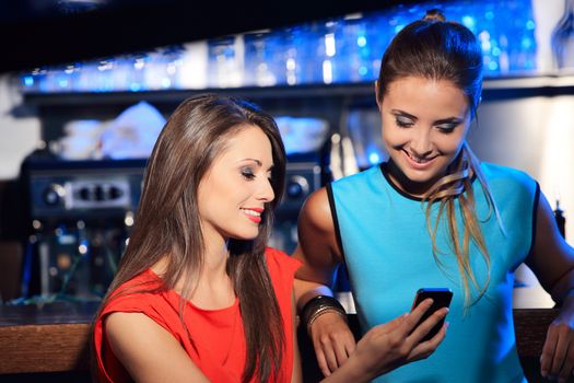 Two beautiful young women friends having fun looking on their smart cell phone