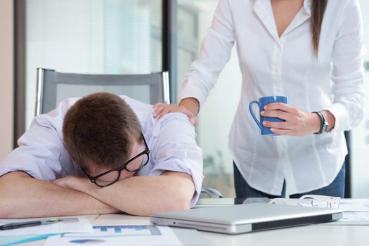 Young office worker is sleeping on the desk, colleague gives him a cup of coffee