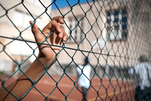 A boy's hand clinging on to a fence, basketball court on background