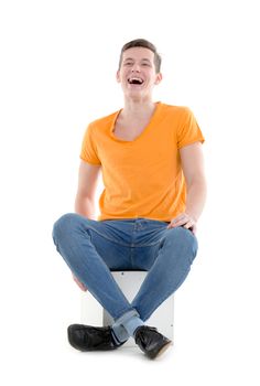 Funny young man, wearing a yellow T-shirt and slim jeans, laughing out loud while sitting on a cube, isolated