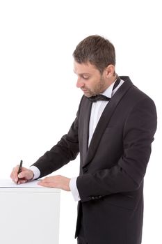 Stylish middle-aged man in a bow tie and suit standing at a pedestal completing a form pausing to read the document with a pen in his hand