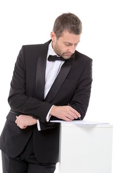 Stylish middle-aged man in a bow tie and suit standing at a pedestal completing a form pausing to read the document with a pen in his hand