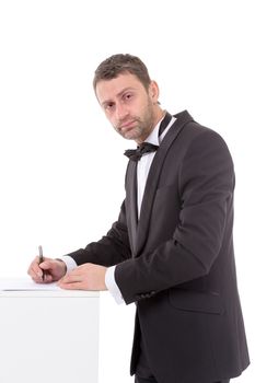 Stylish middle-aged man in a bow tie and suit standing at a pedestal completing a form pausing to read the document with a pen in his hand