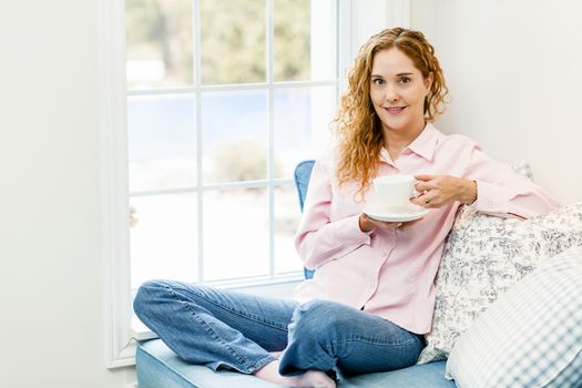 Smiling caucasian woman relaxing by window holding cup of coffee