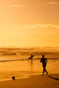 Silhouetted boy playing soccer on beach at sunset with surf boarder in background, Arraial do Cabo, Rio de Janeiro, Brazil.