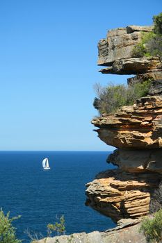 Rocky sandstone cliffs along the coast of Sydney, Australia with a white sailboat in the distance.