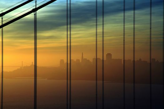 Yellow to blue skies on the early morning on the San Francisco bay area with the Transamerica building silhouette seen through the cables of the Golden Gate bridge.