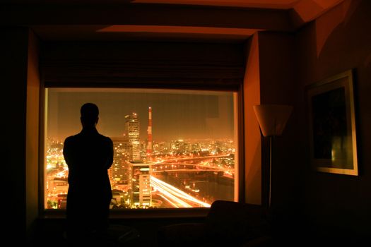 The silhouette of a man with arms crossed in front of a window with a view over a city lit in the night.