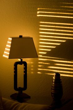 Bright sunshine in a dark room casts lines through window blinds and on to a lamp.