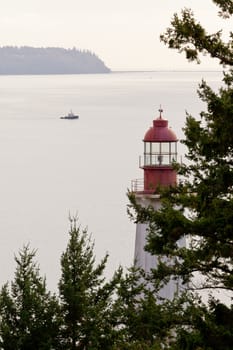 Lighthouse between lush green trees on British Columbia, Canada, West Coast north of Vancouver with small ship out on Pacific ocean