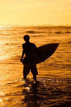 Silhouetted surfer with surfboard in sea at sunset.