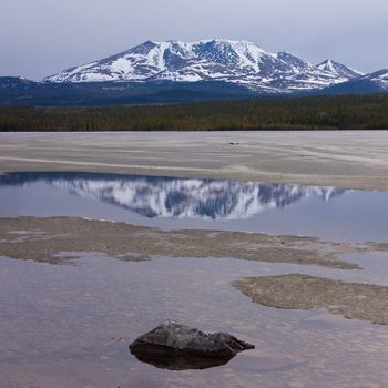 Spring break-up at Fox Lake, Yukon Territory, Canada, still snowy Little Peak reflection on open shore water and rock mimicking a small mountain