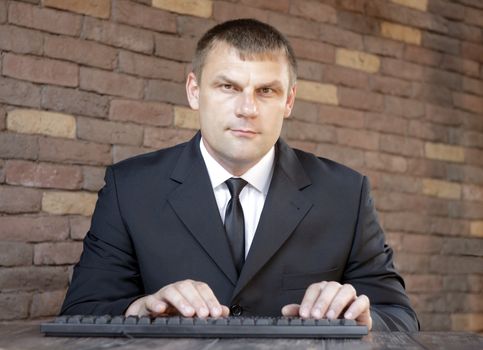 A man is sitting at a wooden table on which the computer keyboard