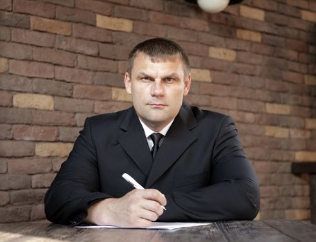 Focused man in a dark suit holding a pen in his hand while sitting at a wooden table against a brick wall