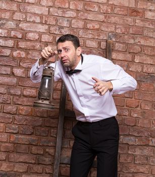 Scared man searching with an oil lamp holding it in front of him as he stands against a brick wall with a terrified anxious expression