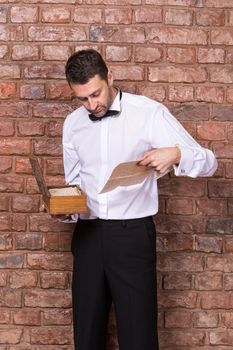 Man standing reading a old document from a wooden box which he is holding in his hand while standing in front of a brick wall