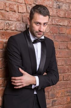 Elegant handsome macho man in a bow tie and tuxedo leaning against a brick wall giving the camera a sultry look