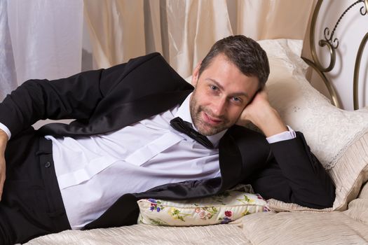 Elegant handsome playboy in a bow tie and suit reclining on a bed in an elaborate bedroom with a seductive smile on his face