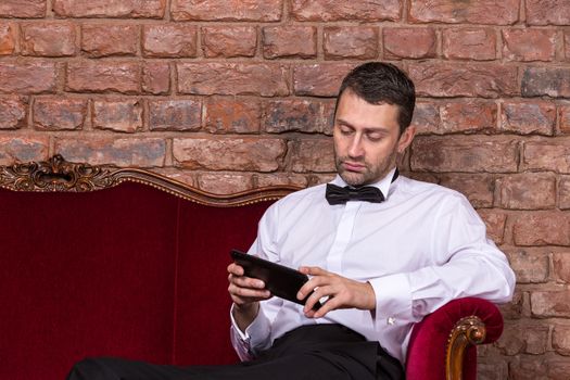 Conceptual image of an elegant businessman lying relaxing on a settee against a brick wall and reading tablet