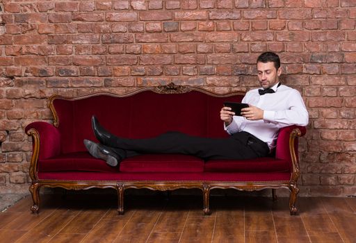 Conceptual image of an elegant businessman lying relaxing on a settee against a brick wall and reading tablet