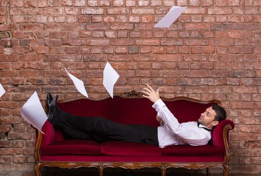 Conceptual image of an elegant businessman lying relaxing on a settee against a brick wall with flying paperwork floating in the air above him