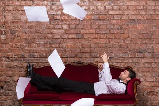 Conceptual image of an elegant businessman lying relaxing on a settee against a brick wall with flying paperwork floating in the air above him