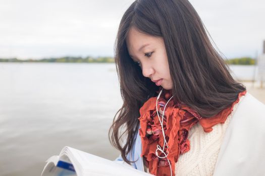 Attractive Asian girl in blue coat reading a book near a lake