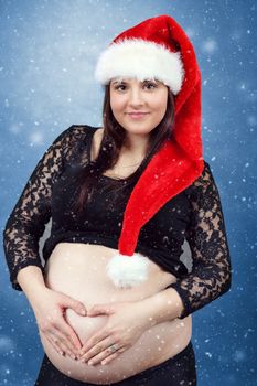 beautiful smiling pregnant young santa woman tenderly holding her tummy, hand in heart shape and with snowing in background