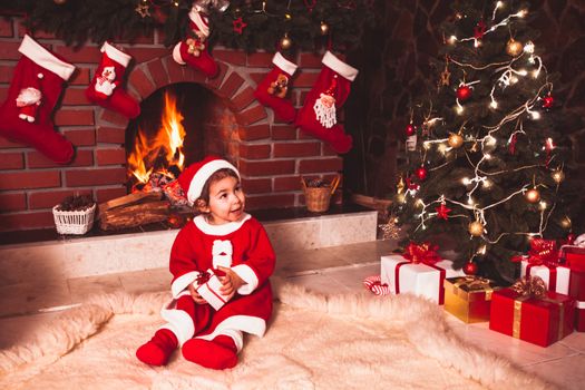 Little girl sitting near fireplace and christmas tree with gift boxes