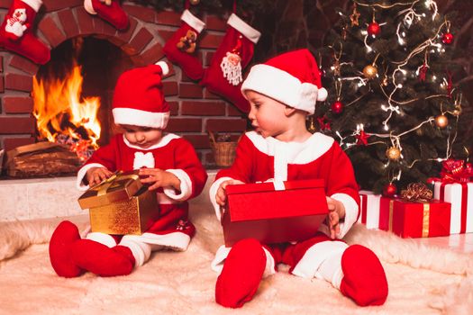 Little girl and boy are sitting near fireplace and christmas tree with gift boxes. Brother and sister in santa suits