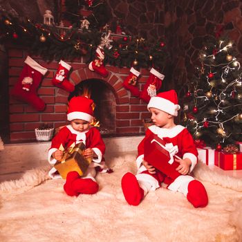 Little girl and boy are sitting near fireplace and christmas tree with gift boxes. Brother and sister in santa suits