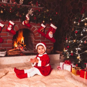 Little girl sitting near fireplace and christmas tree with gift boxes