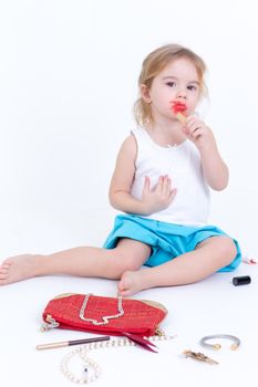 Adorable small girl playing with makeup sitting on the floor with a small bag of cosmetics smearing bright red lipstick on her lips
