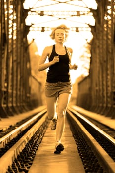 Athlete running on railaway tracks bridge in morning sunrise training for marathon and fitness. Healthy sporty caucasian woman exercising in urban environment before going to work; Active urban lifestyle.