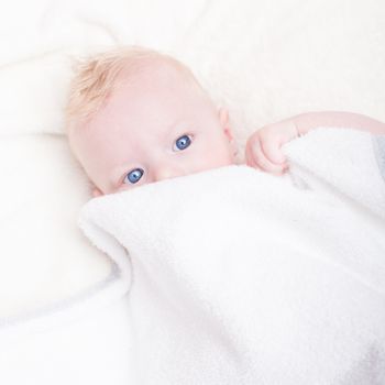 Baby boy with blue eyes hiding under the white blanket.