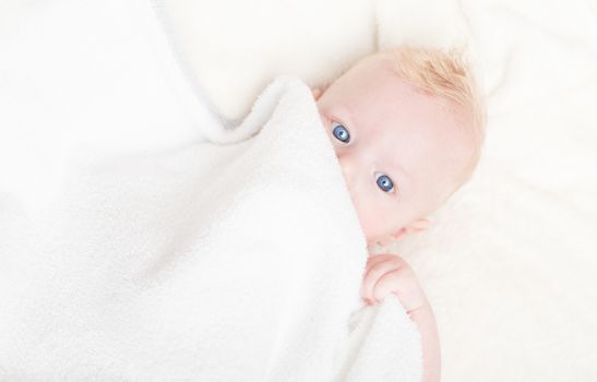 Baby boy with blue eyes hiding under the white blanket.