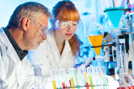 Attractive young female scientist and her senior male supervisor observing color shift of a red liquid in the glass tube in the life science research laboratory (biochemistry, genetics, forensics, microbiology..)